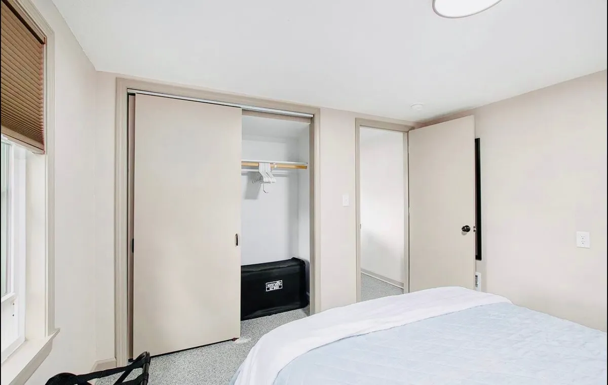 A tranquil bedroom with light blue bedding, an open closet for convenient storage, and neutral-colored walls. Sunlight filters in, highlighting the calm and organized setting meant for restful sleep.