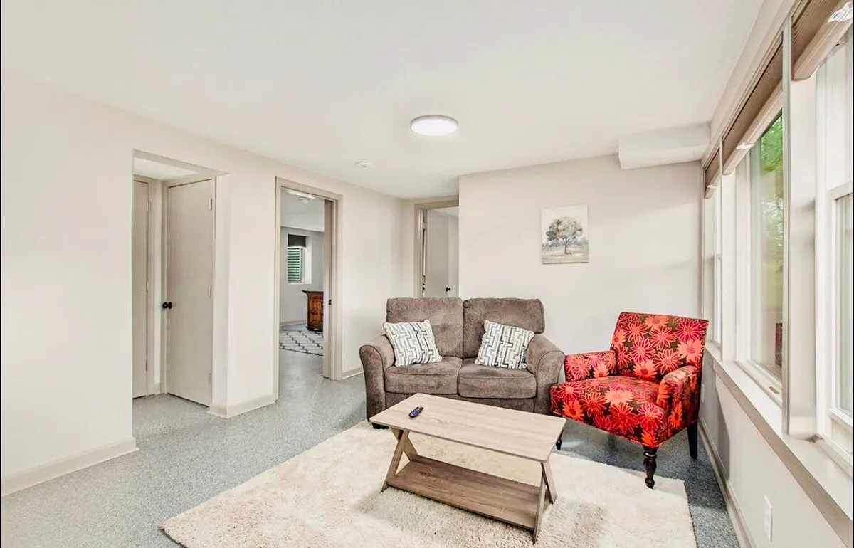A comfortable living space with a brown sofa, red accent chair, coffee table, and an area rug, all bathed in morning sunlight through the windows. The setting invites relaxed conversation or casual reading, offering both warmth and style.