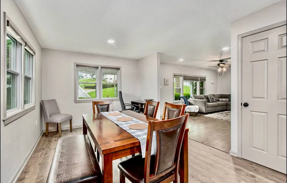 An inviting dining area is shown with a dining table set beside a comfortable living room seating area. Windows offer glimpses of the outdoors, while the arrangement encourages gathering, conversation, and togetherness.