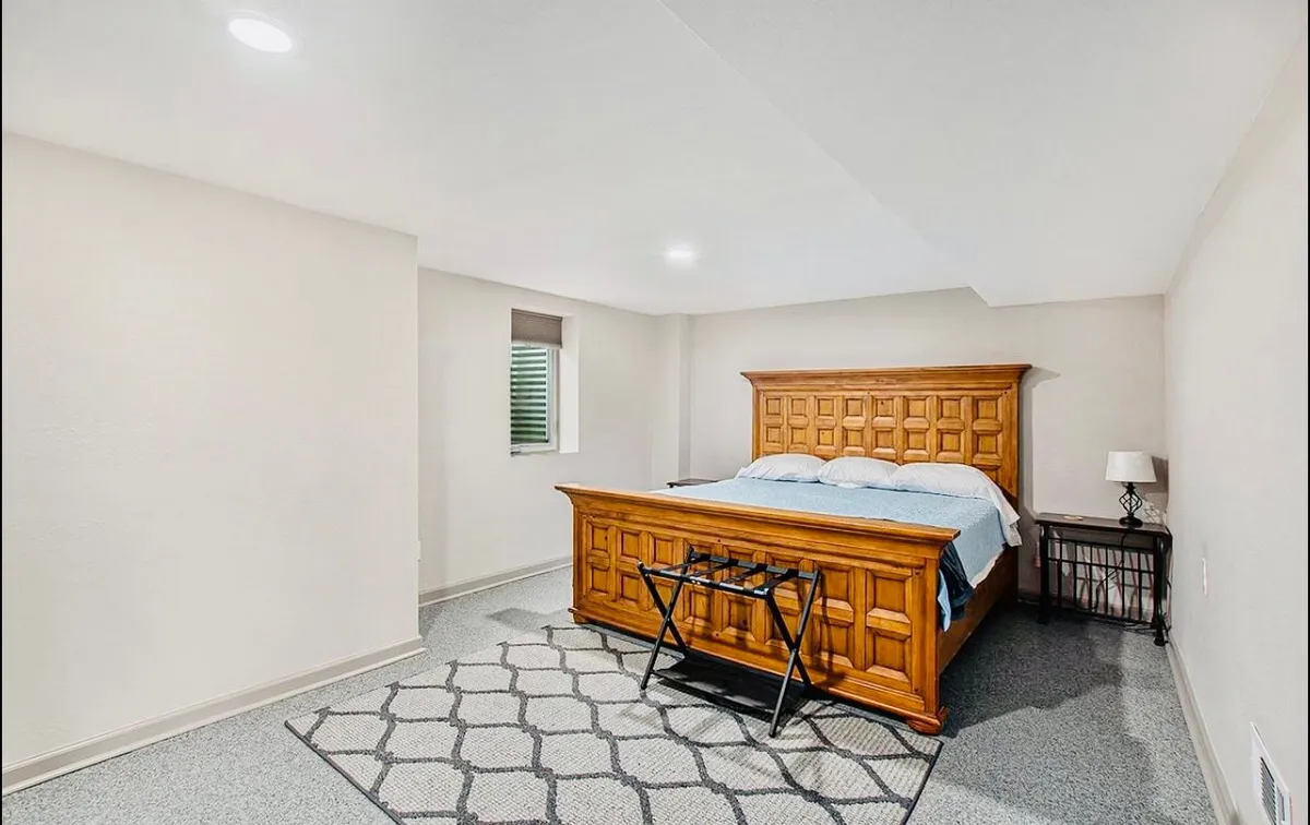 A cozy bedroom featuring a wooden bed frame with light bedding, a decorative rug, and a nightstand with a lamp. Soft natural light streams through the window, creating a peaceful, inviting atmosphere ideal for rest and relaxation.