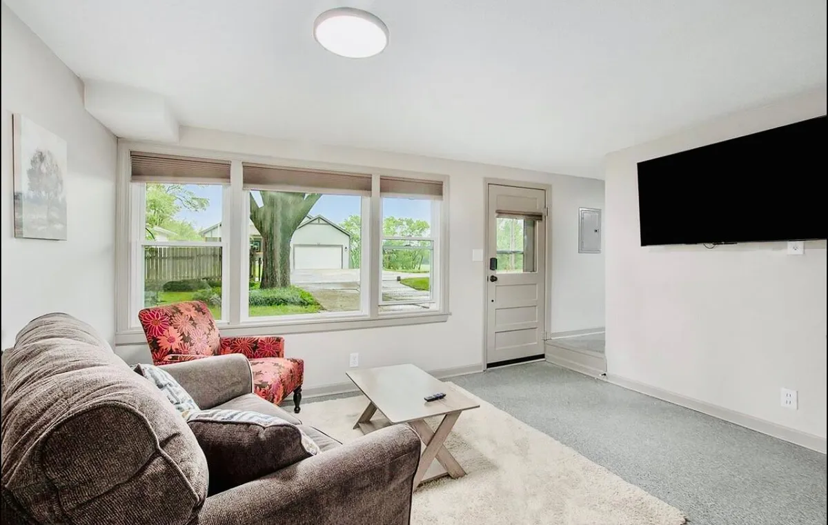 A sun-drenched living room featuring a plush sofa and armchair arranged around a coffee table, with a large TV for entertainment. Expansive windows let in natural light, creating a warm and welcoming atmosphere perfect for relaxing, socializing, or enjoying cozy evenings with blankets and snacks.