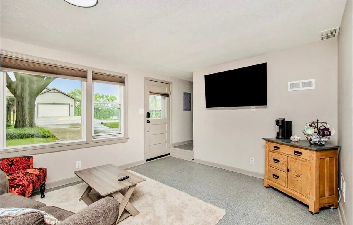 This image shows a welcoming cottage living area featuring a comfortable sofa, a wooden coffee table, large TV, and the entrance door visible in the background. The space exudes warmth and creates a sense of arrival, with soft lighting and natural textures.