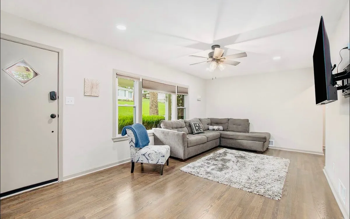 A welcoming living room with a comfortable sofa and chair arranged around a coffee table, facing a television. Large windows let in natural light, creating a bright, airy atmosphere. The room feels perfect for relaxing, entertaining guests, or gathering for a cozy movie night.