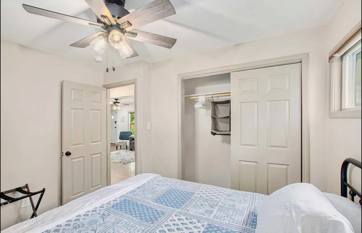 A restful bedroom scene featuring blue patterned bedding, a ceiling fan, and an open closet with clothes hangers, all awash in natural light. The open closet emphasizes functional storage and organization, suitable for visitors with outdoor gear.