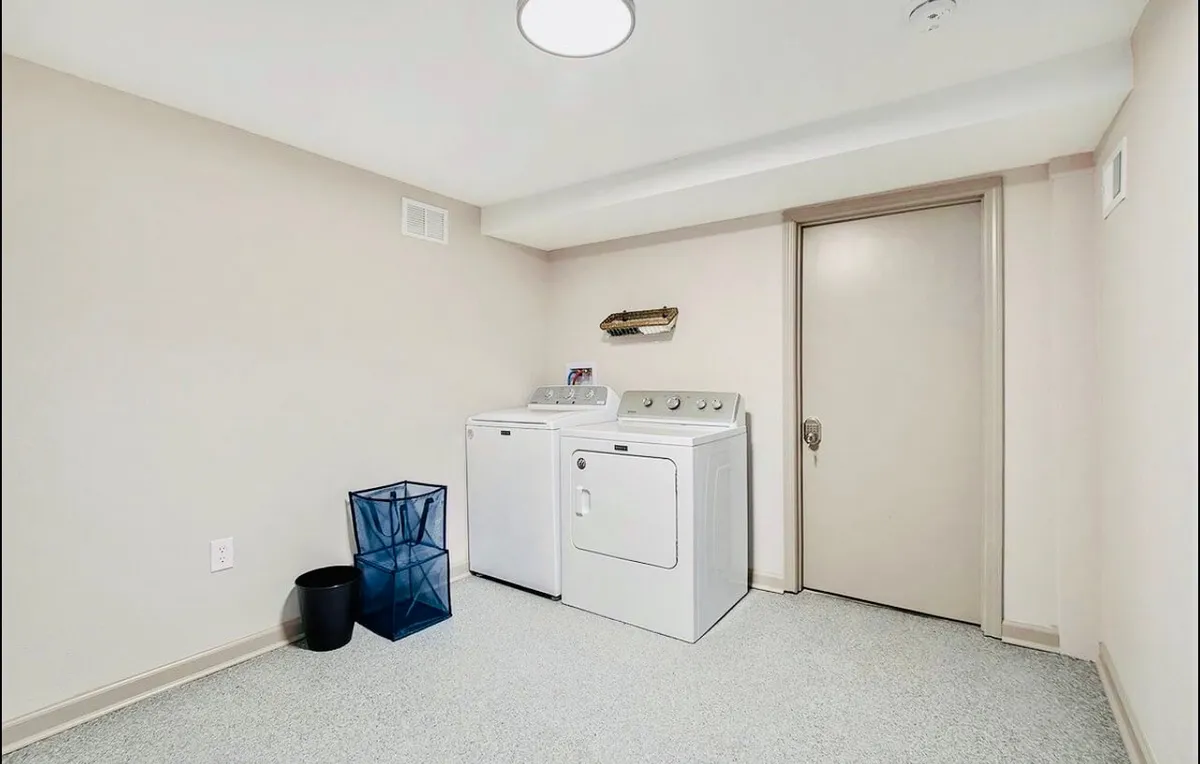 A practical laundry space featuring a washing machine, dryer, blue trash can, and a door. The area is organized, with storage for household items, and conveys a sense of efficient home management.